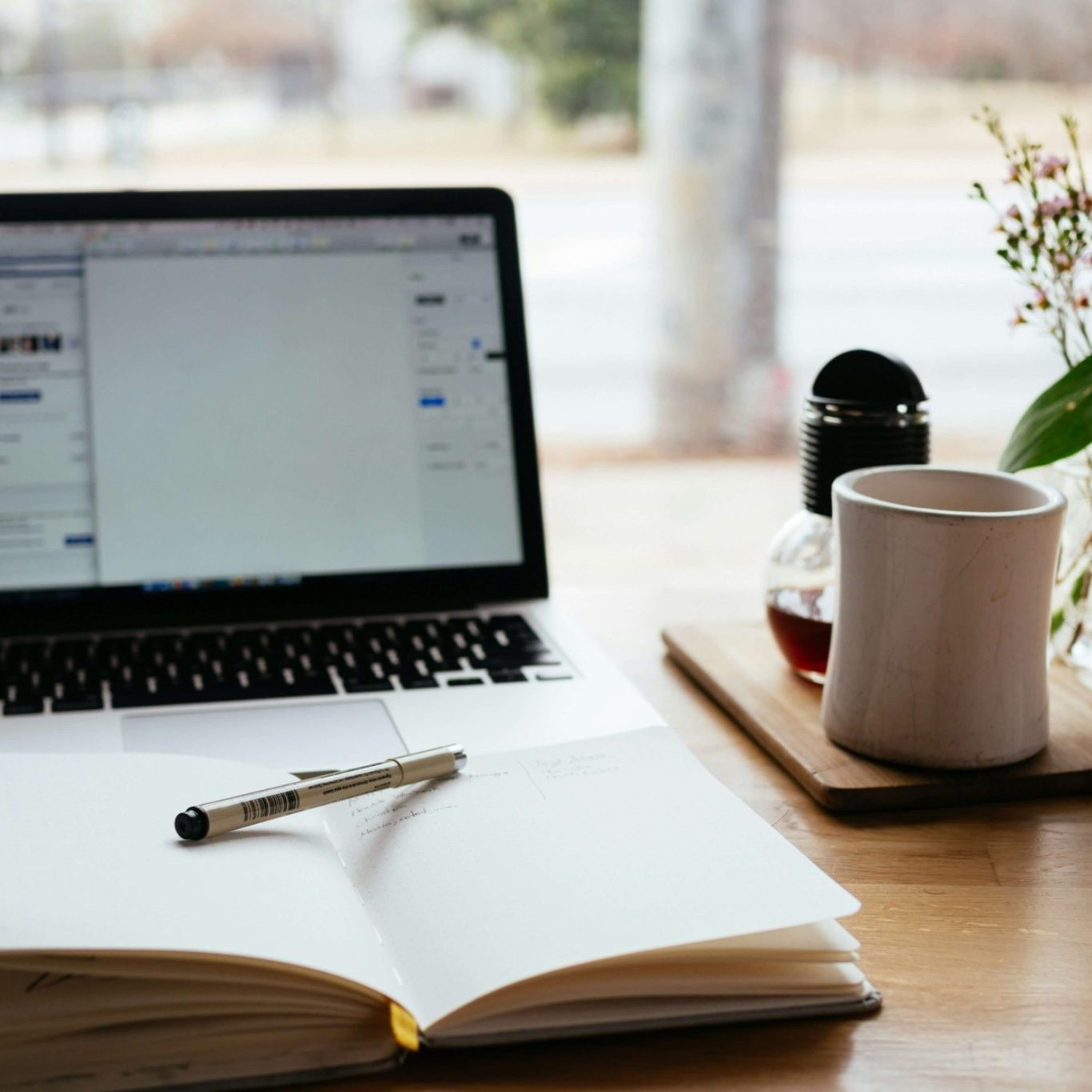 A laptop, an open notebook, a pen, a coffee mug, and a plant on a wooden table.