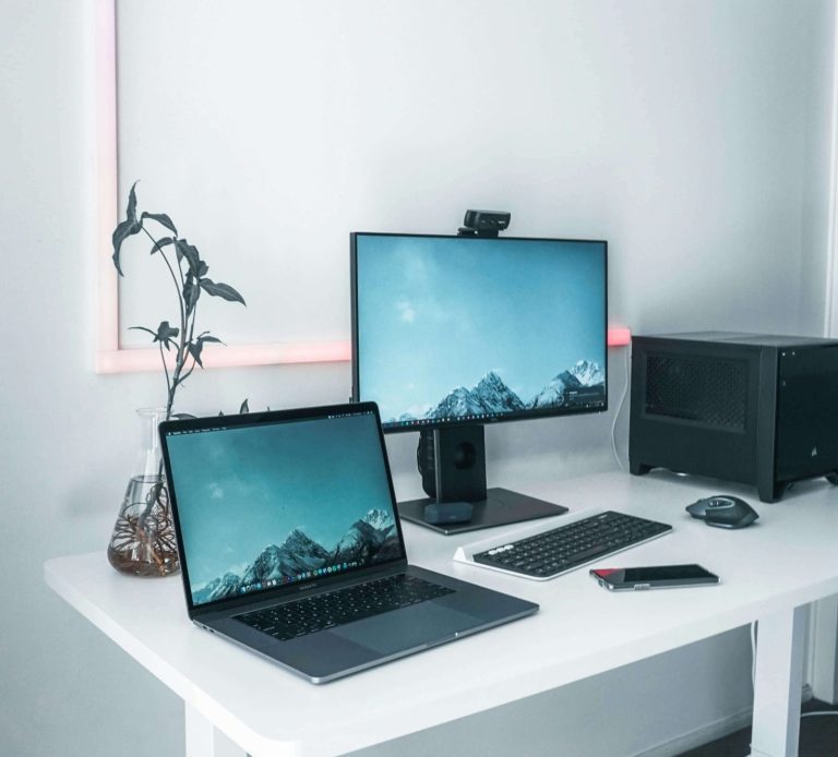 A modern workspace with a laptop, desktop monitor, and potted plant on a white desk.