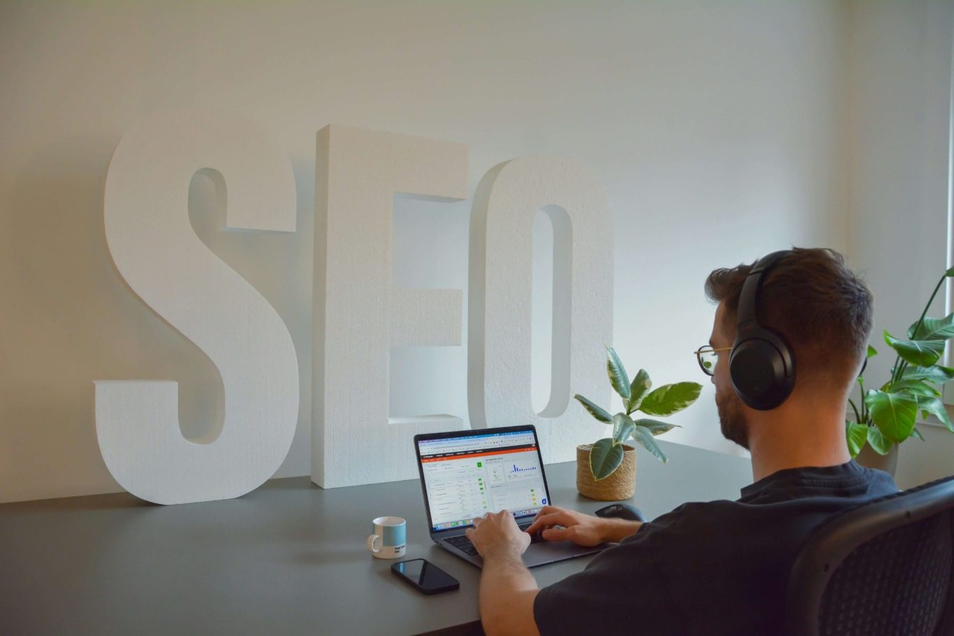 Person wearing headphones working on a laptop with a large "SEO" sign in the background.