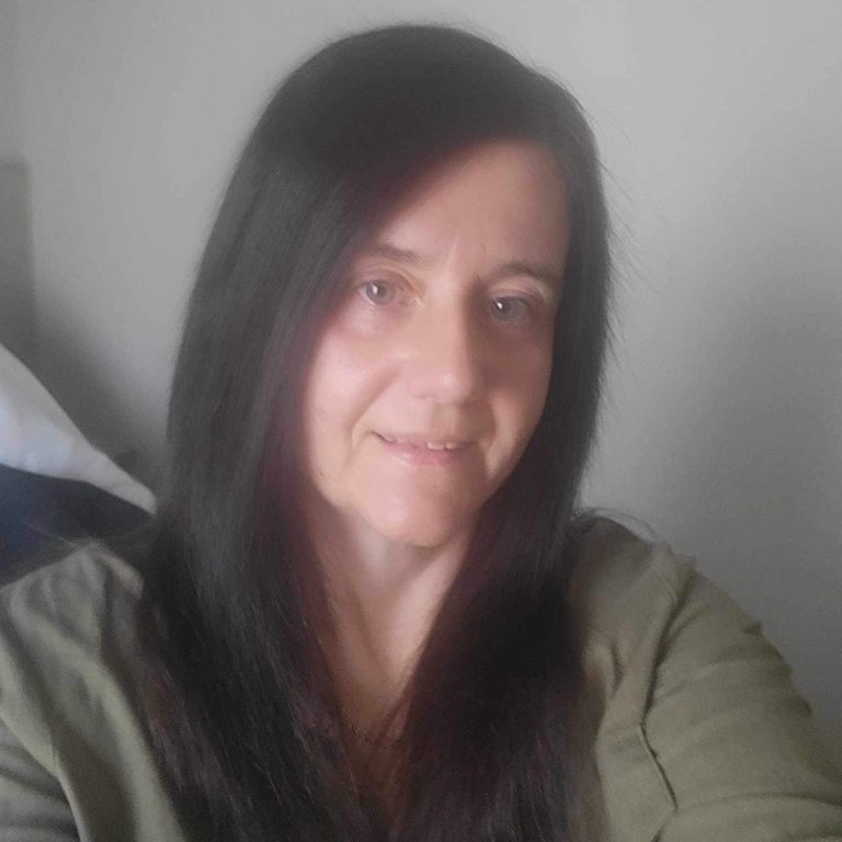 A woman with long black hair smiling, wearing a green top, against a neutral background.