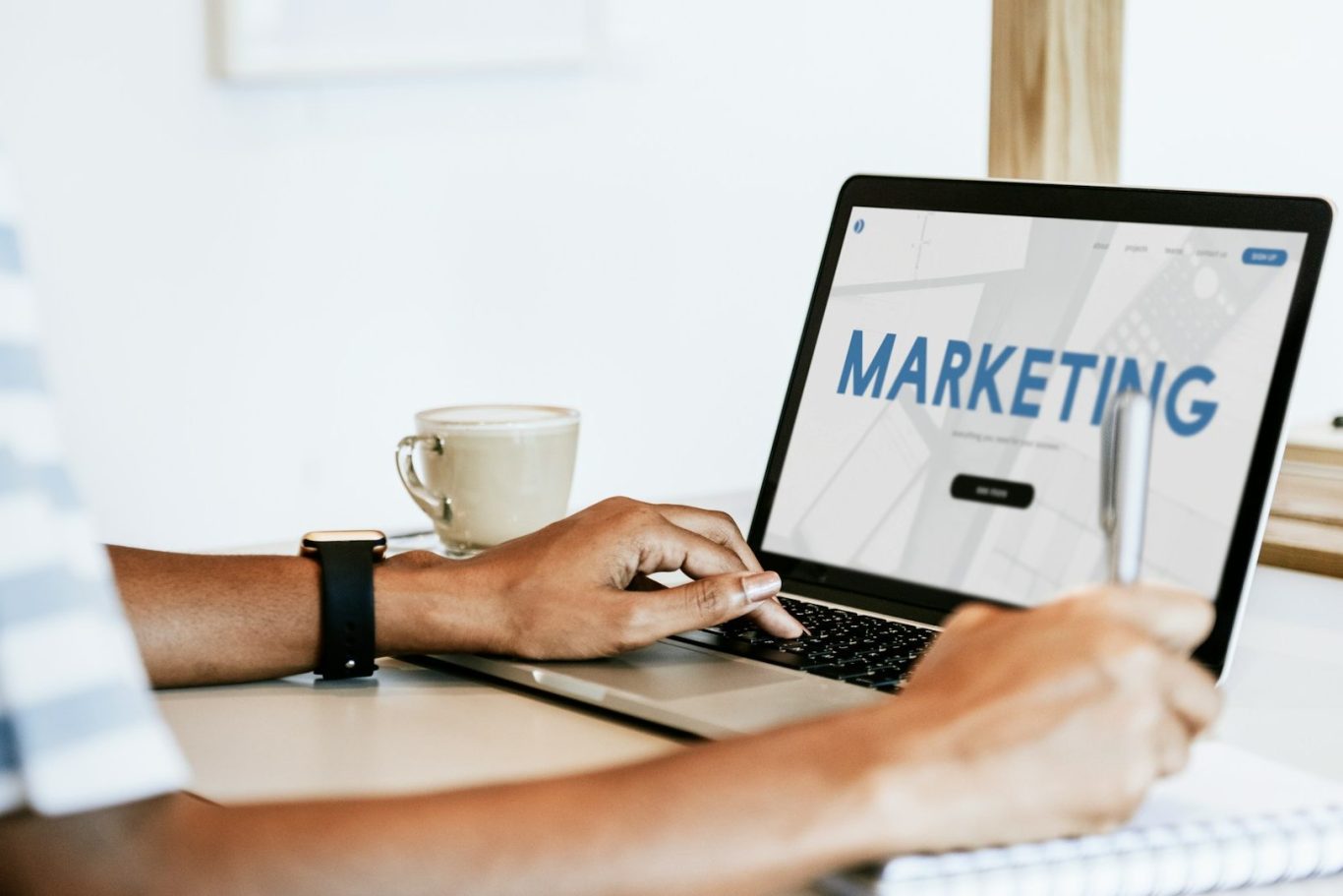Person using a laptop with the word "MARKETING" displayed on the screen, beside a coffee cup.